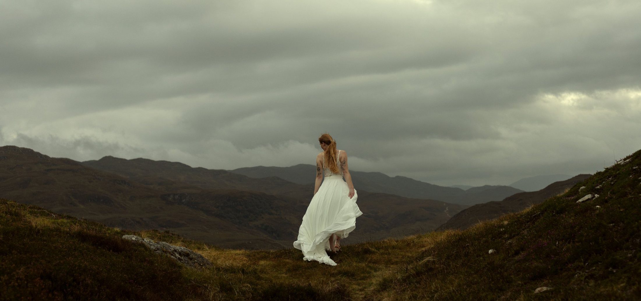 Frau im weißen Kleid geht unter schwerem Wolkenhimmel durch eine Berglandschaft